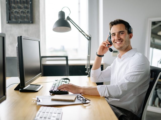 Smiling call center agent working at a desk with a headset and computer in a modern office.